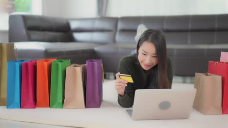 A-young-attractive-woman-lies-on-the-floor-surrounded-by-colorful-shopping-bags-as-she-inputs-her-credit-card-number-to-her-laptop
