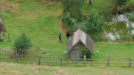 Couple-Walking-By-A-Wooden-Hut-On-The-Lush-Landscape-In-Piaszno,-Pomeranian-Voivodeship,-Poland---high-angle-shot