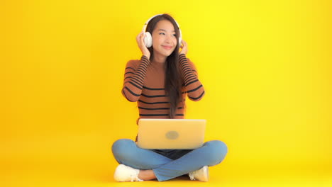 Ein-Hübsches-Asiatisches-Mädchen-Mit-Bluetooth-On-Ear-Kopfhörern,-Das-Auf-Dem-Boden-Sitzt-Und-Musik-Hört,-Die-Einen-Laptop-Auf-Dem-Schoß-Auf-Gelbem-Studiohintergrund-Hält
