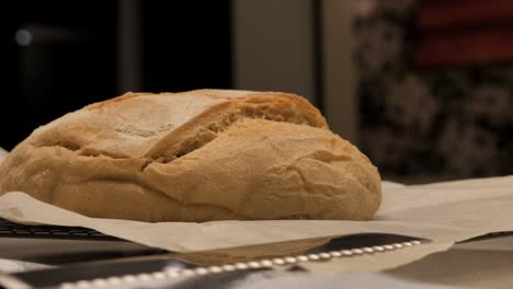 Slow-pan-around-freshly-baked-loaf-of-sour-dough-bread-topped-with-flour-sitting-on-kitchen-bench-with-tea-towel-and-tray,-low-depth-of-field
