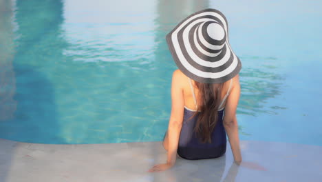 Back-of-Classy-Female-With-Summer-Hat-in-Swimsuit-Sitting-on-Poolside-of-Luxury-Pool