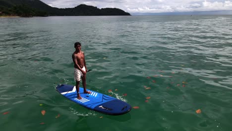 Aerial-drone-pivot-circular-bird's-eye-view-of-man-exercising-on-a-sup-paddle-board-in-turquoise-tropical-clear-waters,-with-beach-jungle-and-coastline-in-Thailand