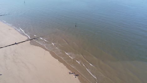 Sunny-aerial-view-looking-down-over-golden-sandy-summer-beach-coastal-tide-with-tourists-walking-below