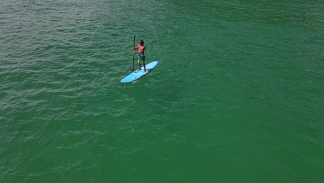 Aerial-drone-bird's-eye-view-of-Caucasian-man-exercising-on-a-sup-paddle-board-in-turquoise-tropical-clear-waters,-with-beach-and-coastline-in-Thailand