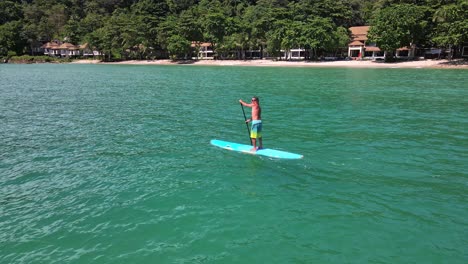 Low-angle-aerial-drone-bird's-eye-view-of-Caucasian-man-exercising-on-a-stand-up-paddle-board-in-turquoise-tropical-clear-waters,-with-beach,-resort-and-coastline-in-Thailand