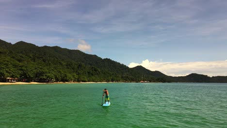 low-angle-aerial-drone-bird's-eye-view-of-Caucasian-man-exercising-on-a-sup-paddle-board-in-turquoise-tropical-clear-waters,-with-beach-and-coastline-in-Thailand