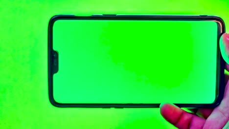 Nahaufnahmehand-Eines-Asiatischen-Mannes,-Der-Ein-Modernes-Smartphone-Vor-Grünem-Hintergrund-Mit-Grünem-Display-Hält
