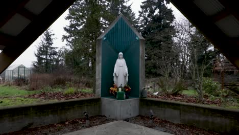 Statue-Der-Jungfrau-Maria-In-Einem-Altar-Vor-Einer-Kleinen-Holzkapelle-Im-üppigen-Park-Ohne-Menschen---Drohnenrückzugsaufnahme