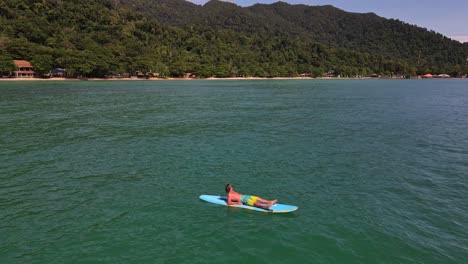 retreat-reveal-aerial-drone-bird's-eye-view-of-Caucasian-man-relaxing-and-lying-down-on-a-stand-up-paddle-board-in-turquoise-tropical-clear-waters,-with-beach-and-coastline-in-Thailand