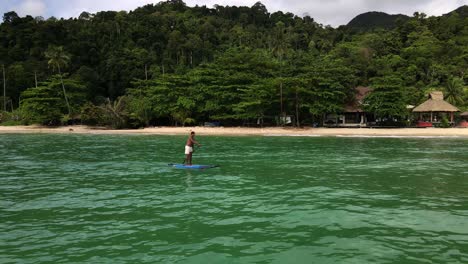 low-angel-Aerial-drone-tracking-shot-of-Asian-man-exercising-on-a-sup-paddle-board-in-turquoise-tropical-clear-waters,-with-beach,-small-bungalow-beach-resort-and-coastline-in-Thailand
