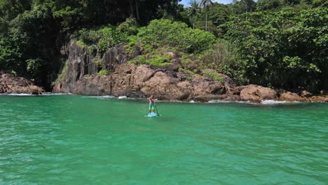 low-angle-Aerial-drone-bird's-eye-view-of-man-exercising-on-a-sup-paddle-board-in-turquoise-tropical-clear-waters,-rocky-coastline-and-tropical-jungle-forest-in-Thailand