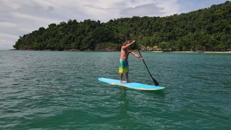 low-angle-backwards-dolly-shot-drone-view-of-Caucasian-man-exercising-on-a-sup-paddle-board-in-turquoise-tropical-clear-waters,-with-beach-and-coastline-in-Thailand