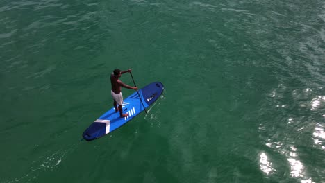 Aerial-drone-bird's-eye-view-of-Asian-man-exercising-on-a-sup-paddle-board-in-turquoise-tropical-clear-waters
