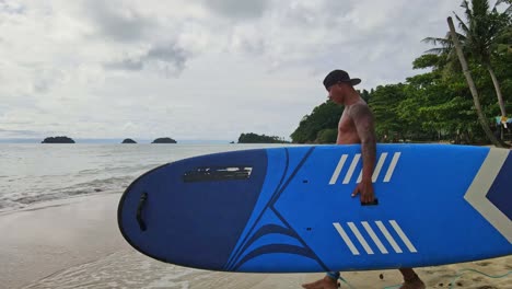 Asian-man-walking-with-a-sup-paddle-board-on-a-beach-towards-the-ocean-with-Islands-and-coastline-in-Thailand
