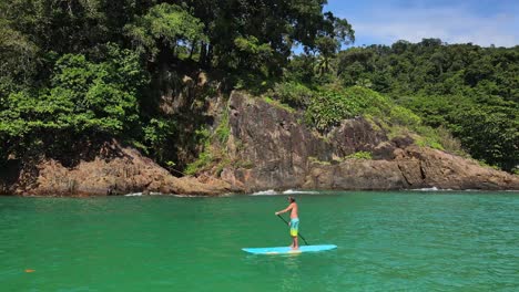 Trucking-aerial-drone-shot-of-Caucasian-man-exercising-on-a-sup-paddle-board-in-turquoise-tropical-clear-waters,-with-jungle-tropical-coastline-in-Thailand