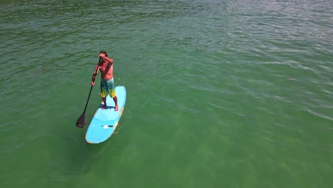static-aerial-drone-shot-of-Caucasian-man-exercising-on-a-sup-paddle-board-approaches-camera-in-turquoise-tropical-clear-waters