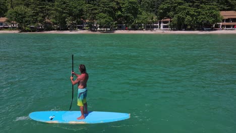 Low-angle-trucking-Aerial-drone-bird's-eye-view-of-Caucasian-man-exercising-on-a-sup-paddle-board-in-turquoise-tropical-clear-waters,-with-beach-resort-and-coastline-in-Thailand