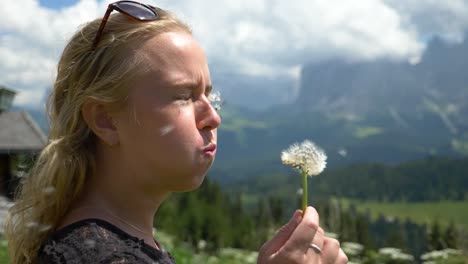 Blonde-Female-Model-blowing-a-Dandelion-flower-in-the-wind-the-wrong-way-in-the-Val-Gardena-area,-Dolomites-Italy