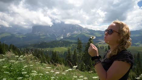 Blonde-girl-blowing-a-dandelion-flower-in-the-wind,-beautiful-mountains-nature-setting-sunny-summer-day-at-Seiser-Alm,-Val-Gardena,-Dolomiti