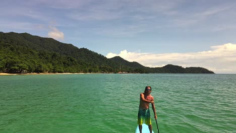 static-low-angle-aerial-drone-shot-of-Caucasian-man-exercising-on-a-sup-paddle-board-in-turquoise-tropical-clear-waters,-with-beach-and-coastline-in-Thailand