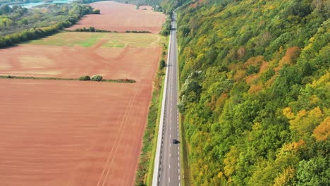 Aerial-shot-of-the-car-riding-straight-the-road-in-autumn,-sunny-day-with-colorful-forest,-going-away