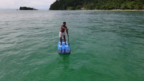 Aerial-drone-low-angle-of-Asian-man-exercising-on-a-sup-paddle-board-in-turquoise-tropical-clear-waters,-with-beach,-Islands-and-coastline-in-Thailand
