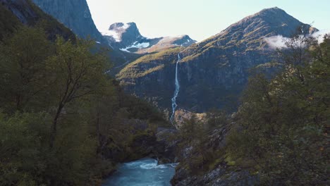 Die-Kamera-Schwenkt-Langsam-Durch-Einen-Offenen-Wald-Mit-Einem-Wunderschönen-Fluss-Und-Einem-Wasserfall,-Der-Im-Hintergrund-Einen-Berg-Hinunterkommt