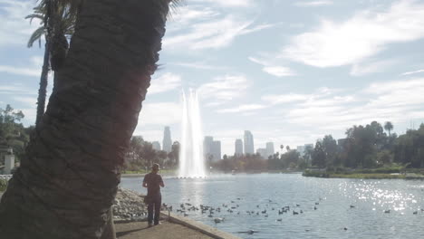 Single-Man-Goes-Fishing-Standing-Up-at-Echo-Park-Lake,-with-Downtown-LA-City-in-Background