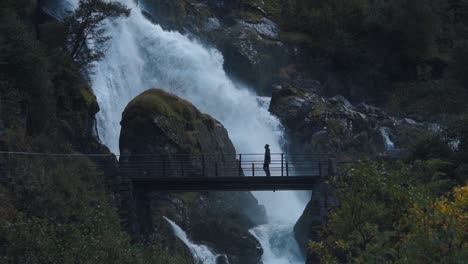 Slow-motion-shot-of-man-walking-infront-of-a-wild-waterfall-over-a-small-wooden-bridge,-Norway-Briksdalen