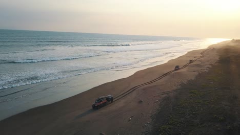 Drone-aerial-of-4x4-Cars-driving-on-the-shore-of-black-sand-beach-during-sunset