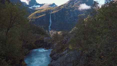 Drone-panning-over-a-small-creek,-flying-up-and-revealing-beautiful-scenic-view-of-a-waterfall-coming-down-from-tall-mountains