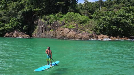 backwards-dolly-aerial-drone-shot-of-Caucasian-man-exercising-on-a-sup-paddle-board-in-turquoise-tropical-clear-waters,-with-tropical-jungle-palm-trees-and-coastline-in-Thailand