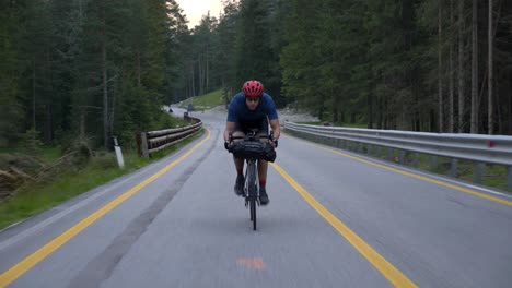 Ciclista-Con-Casco-Rojo-En-Bicicleta-Por-Carretera-Asfaltada-En-Las-Montañas-Con-Líneas-Amarillas-En-Un-Bosque-De-Pinos,-Dolomitas,-Italia