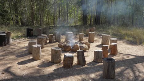 Tree-Stumps-Around-Firepit-With-Smoke-Rising-From-The-Burning-Logs---Camping-Area-In-Mount-Byron,-Queensland,-Australia