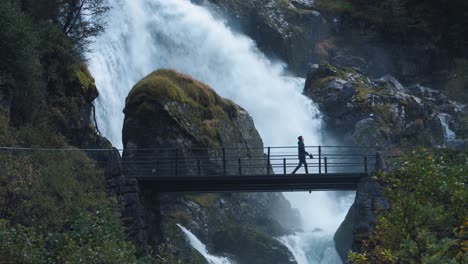 Man-walking-across-a-small-bridge-with-a-wild-waterfall-on-the-background,-Norway-Briksdalen