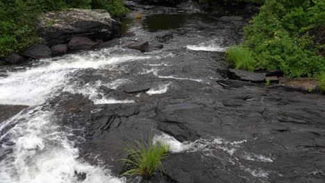 Rocky-River-Splashing-Downstream---White-Water-Creek-Over-Black-Rocks-Surrounded-by-Green-Vegetation