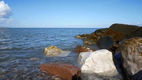 Sunny-blue-ocean-tide-washing-up-onto-colourful-stone-pebble-beach-coastline-island-holiday-scene-dolly-right