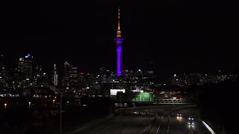 Fahrzeuge,-Die-Während-Der-Pandemie-Des-Coronavirus-In-Auckland,-Neuseeland,-Auf-Der-Stadtstraße-Mit-Nachts-Beleuchtetem-Himmelsturm-Und-Skyline-Fahren