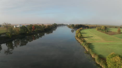 Serene-calm-morning-with-early-autumn-color-on-trees-seen-from-drone-flying-over-calm-river-flowing-through-farm-fields