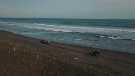 Amazing-drone-aerial-shot-of-4x4-cars-driving-over-black-sand,-waves-crashing,-camping-trip-in-San-Jose,-Guatemala