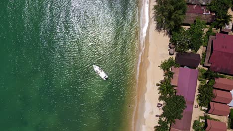 Tropical-Island-drone,-bird’s-eye-view-down-shot-with-lush-green-rain-forest-and-tropical-palm-trees-with-small-bungalow-resort-on-a-white-sand-beach-with-speed-boat-in-water