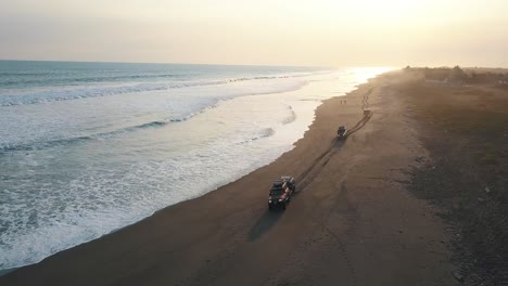 Drone-aerial-shot-of-3-cars-driving-over-the-sand,-waves-crashing,-shore-in-San-Jose,-Guatemala