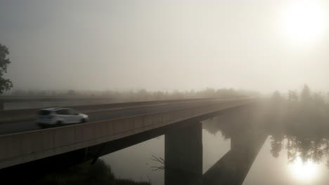 Ascending-aerial-view-of-cars-on-highway-bridge-crossing-calm-river-as-morning-light-shines-through-dense-fog-hanging-over-the-landscape