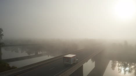 Dangerous-foggy-driving-conditions-with-cars-and-trucks-driving-over-highway-bridge-crossing-river-with-rising-sun-and-dense-fog