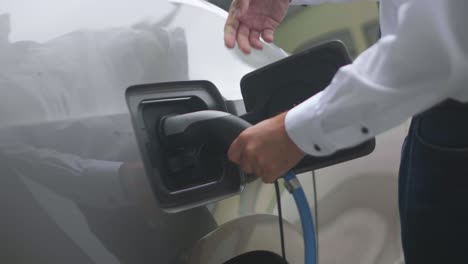 Close-up-of-a-modern-electric-car-charger-being-attached-to-a-silver-electric-car-by-a-person-wearing-a-white-shirt