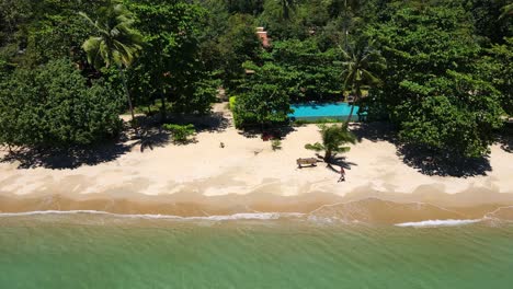 Tropical-Island-drone,-bird’s-eye-view-down-shot-with-lush-green-rain-forest-and-tropical-palm-trees-with-white-sand-beach-with-alone-man-walking-past-a-resort-on-the-beach-with-no-tourists