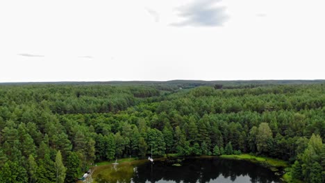 Drone-Descending-Towards-A-Clear-Lake-With-Mooring-Boats-In-The-Middle-Of-Dense-Pine-Forest-Landscape-In-Pradzonka-In-Northern-Poland---Wide-Shot