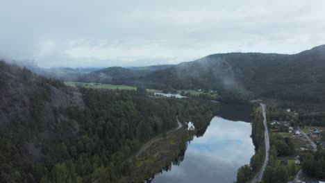Aerial-drone-view-of-a-reflecting-river,-town-and-hills,-dark,-foggy-day,-in-Aamli,-South-Norway