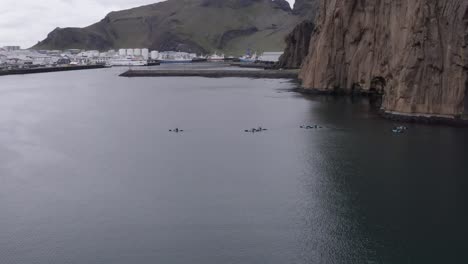 Kayakers-crossing-bay-near-harbor-of-Vestmannaeyjar-with-rocky-cliffs,-aerial