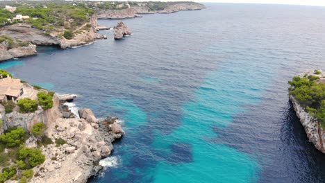 Aerial-over-the-crystal-clear-blue-waters-of-Mallorca-island-surrounded-by-rocky-cliffs-in-Spain